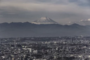 A sprawling cityscape in the foreground with a snow-capped Mount Fuji rising above a mountain range in the distance.