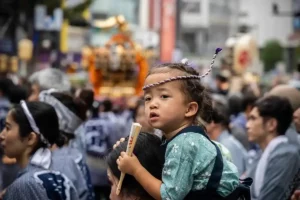 A young child in a green happi coat and rope headband sits on an adult's shoulders, watching a crowded Japanese festival.