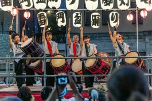 A group of Japanese taiko drummers performs energetically on a stage at night under large, illuminated paper lanterns for a crowd.