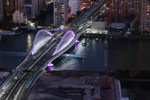 High-angle night shot of a glowing double-arch bridge, with traffic crossing and a boat passing on the dark river below.