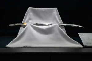 A polished Japanese katana blade resting on a white draped stand in a dark museum-style display.