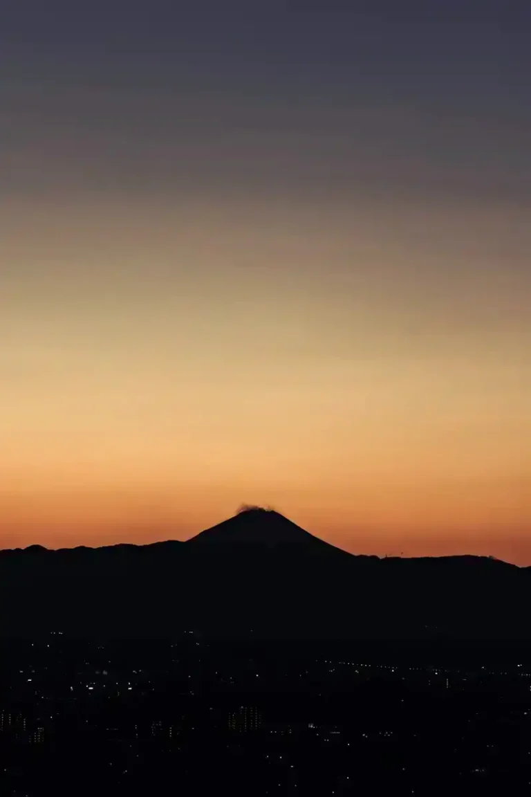 Silhouetted Mount Fuji against a vibrant orange and purple sunset sky, overlooking distant city lights at dusk.